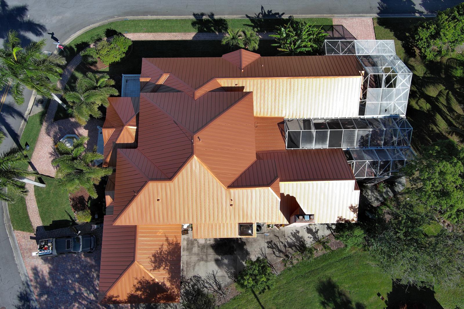Top-down drone view of a Florida home with a copper standing seam metal roof, highlighting multiple roof sections, screened pool enclosures, driveway, and surrounding landscaping.