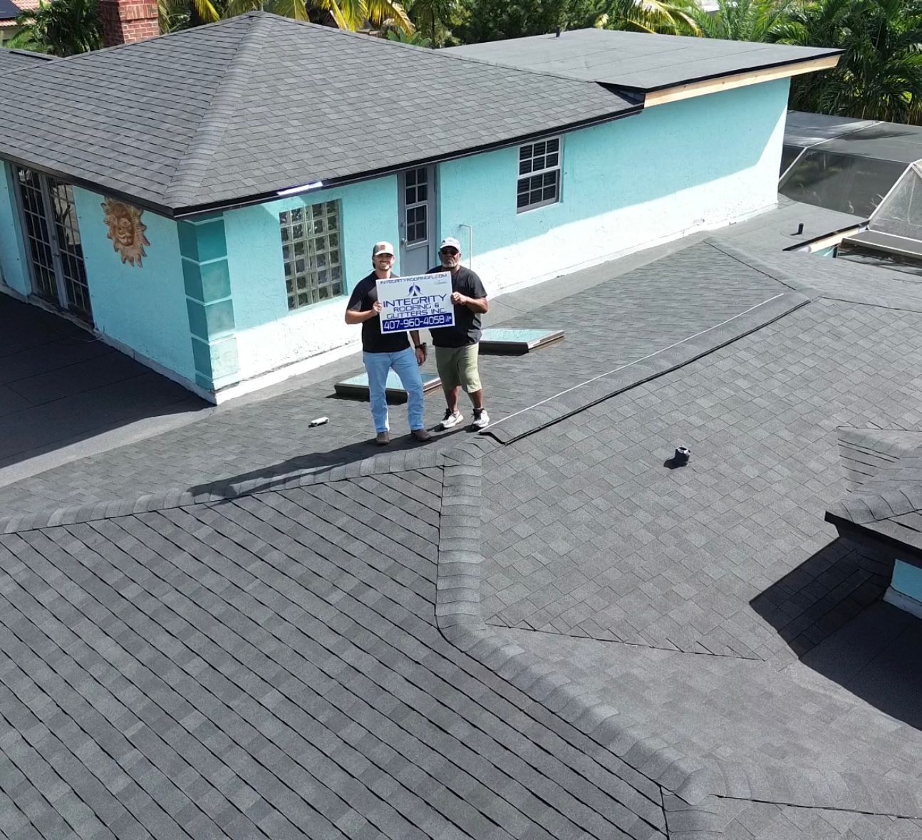 Aerial drone photo of two roofing professionals standing on a newly installed gray shingle roof, holding a company sign, with a turquoise single-story home and surrounding palm trees visible in the background.
