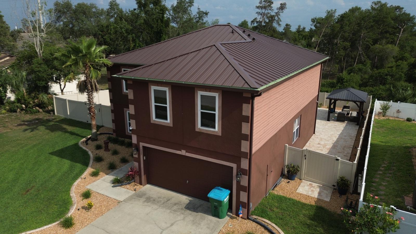 "Residential home with a brown metal roof, surrounded by lush greenery and a well-maintained yard. The image showcases a metal roof replacement project, highlighting its modern design and durability for Florida homes.