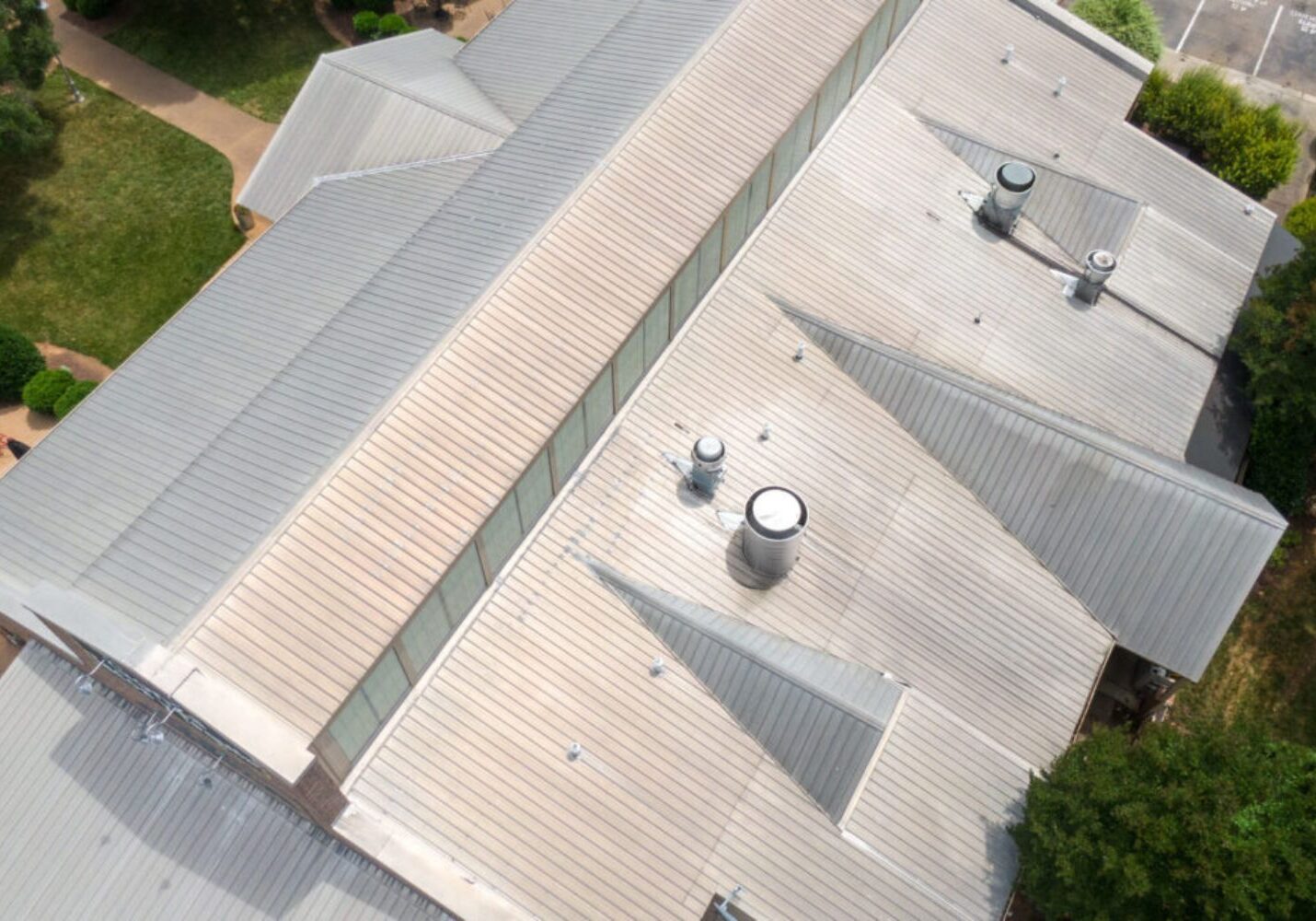 Drone view of a large standing-seam metal roof with multiple roof sections and vents, on a commercial-style building surrounded by trees and pavement.