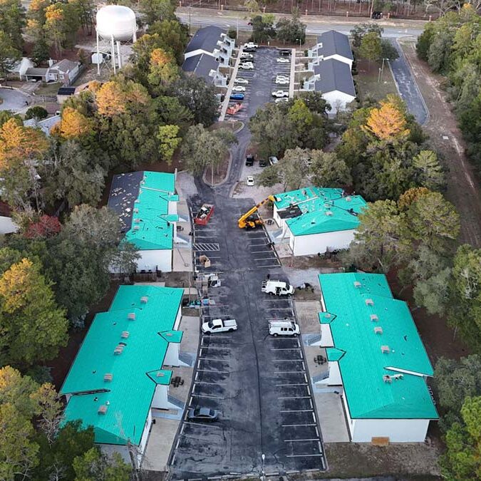 Aerial view of a small apartment or townhouse complex surrounded by trees, featuring multiple low-rise buildings with teal roofing under installation, parked work vehicles, construction equipment, and a central paved driveway with parking spaces.