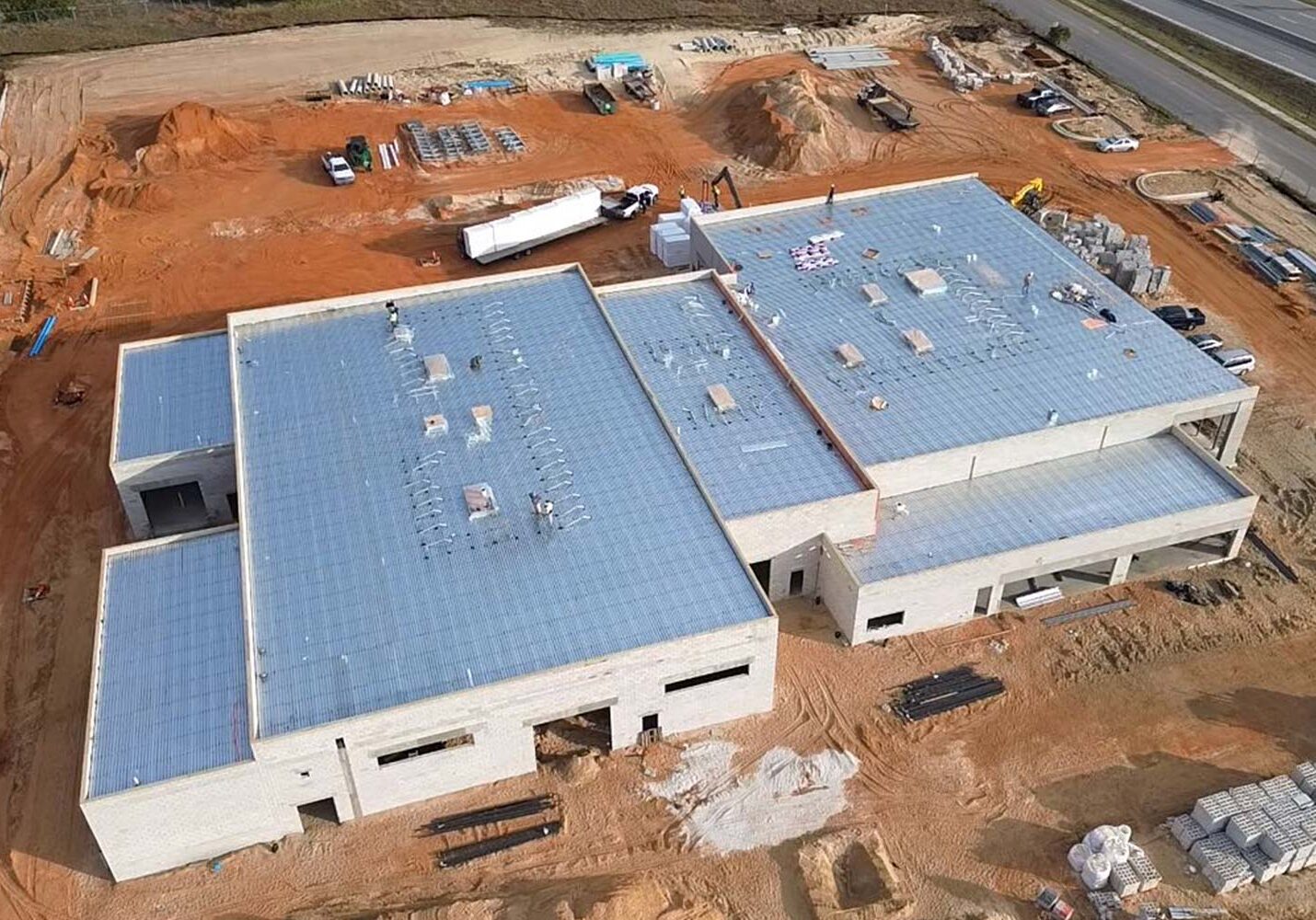 Commercial project in progress with a light‑colored flat roof, construction materials, trucks, and graded soil around the site.