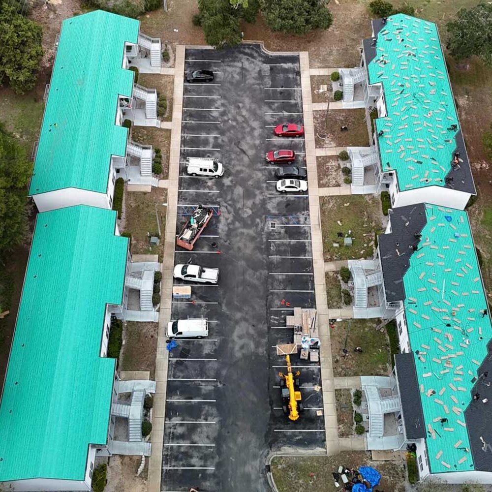erial view of multi-unit residential buildings with teal underlayment on roofs during replacement work, showing construction vehicles and parked cars in the driveway.