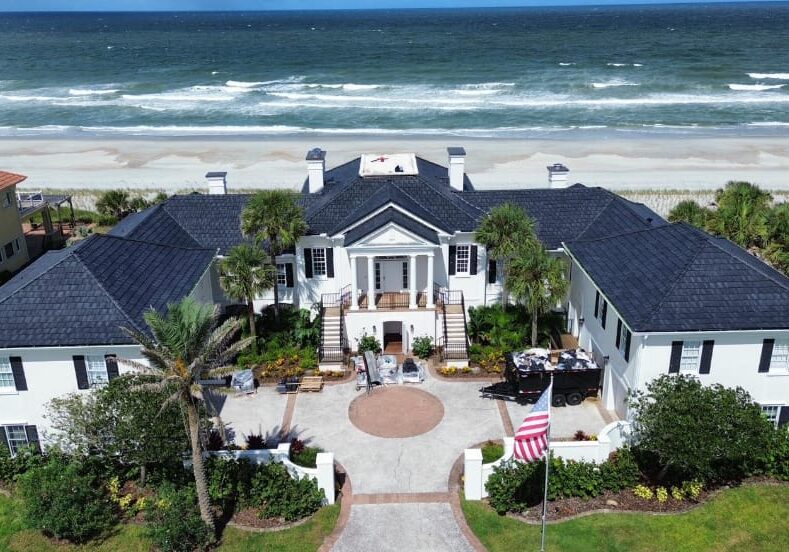 Aerial drone view of a coastal Florida luxury home with a newly installed dark gray asphalt shingle roof, showing symmetrical rooflines, oceanfront setting, and landscaped grounds.
