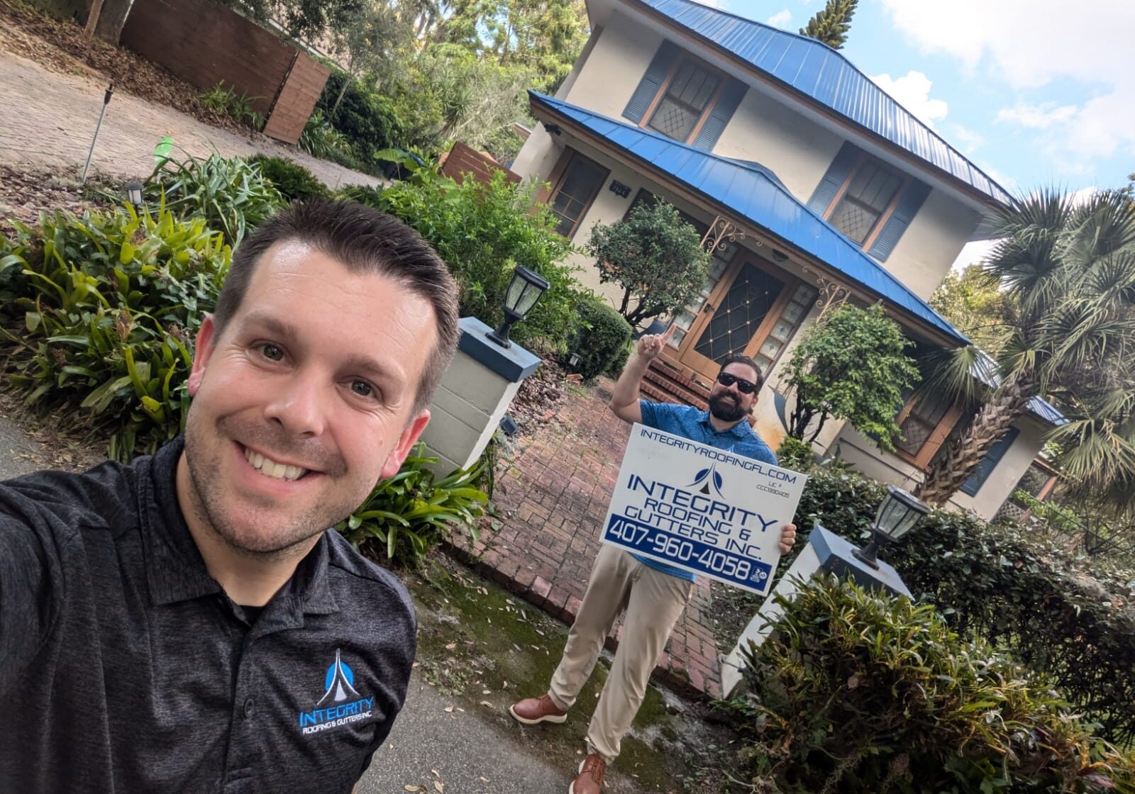 Integrity Roofing & Gutters team members standing in front of a Florida home with a newly installed blue metal roof, holding a company sign after completing a residential roof replacement.