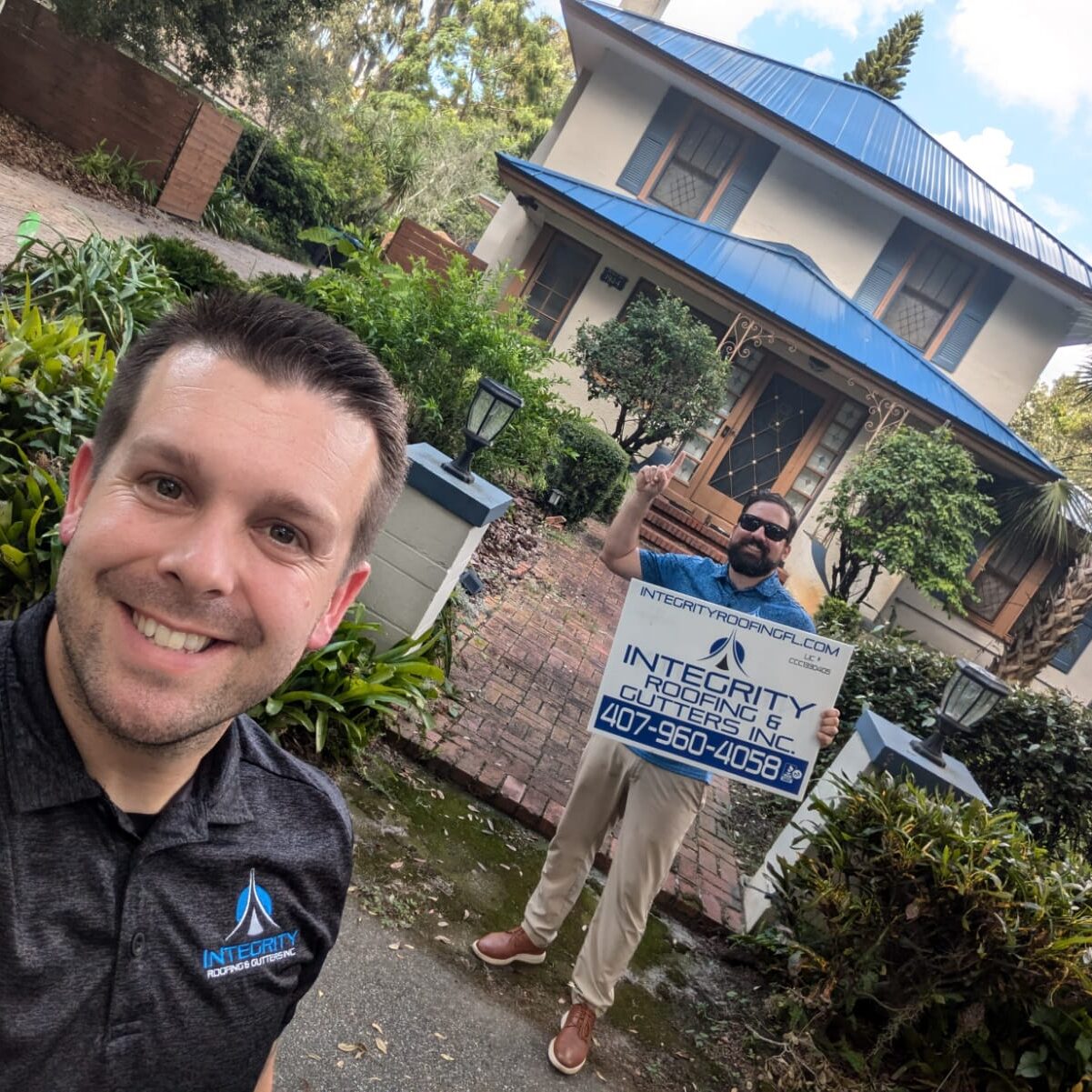 Integrity Roofing & Gutters team members standing in front of a Florida home with a newly installed blue metal roof, holding a company sign after completing a residential roof replacement.