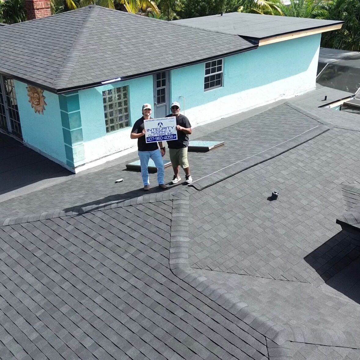 Aerial drone photo of two roofing professionals standing on a newly installed gray shingle roof, holding a company sign, with a turquoise single-story home and surrounding palm trees visible in the background.