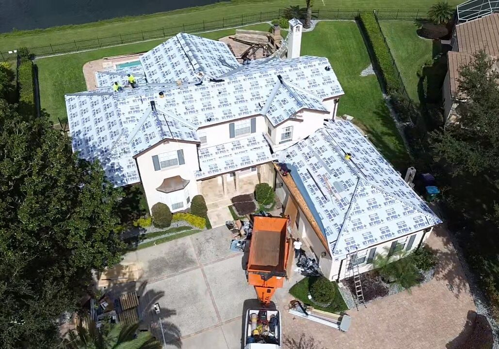 Aerial drone view of a large residential home undergoing roof replacement, with protective underlayment installed across multiple roof sections, roofing crew working on the roof, a debris container in the driveway, and a landscaped backyard near a waterfront.